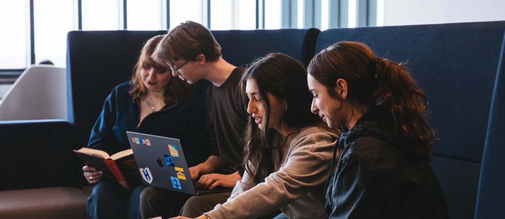 Four young adults sit together on a dark blue couch. Two read a book and a laptop with stickers, while the other two look at papers and a laptop, appearing focused and engaged in group work or study.