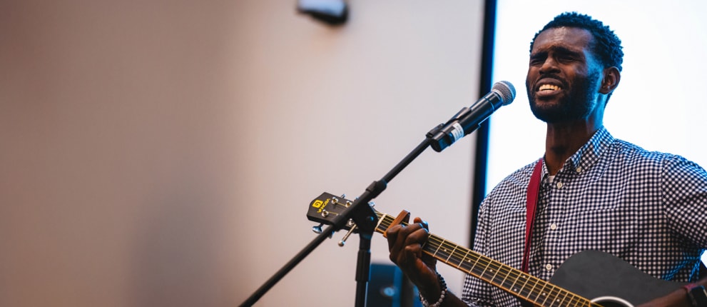 A man in a checkered shirt sings passionately into a microphone while playing an acoustic guitar on stage.