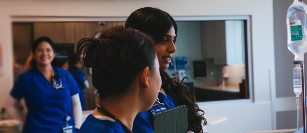 Three healthcare professionals in blue scrubs are in a medical setting. Two are in the foreground having a conversation, while one smiles in the background. An IV stand is visible on the right.