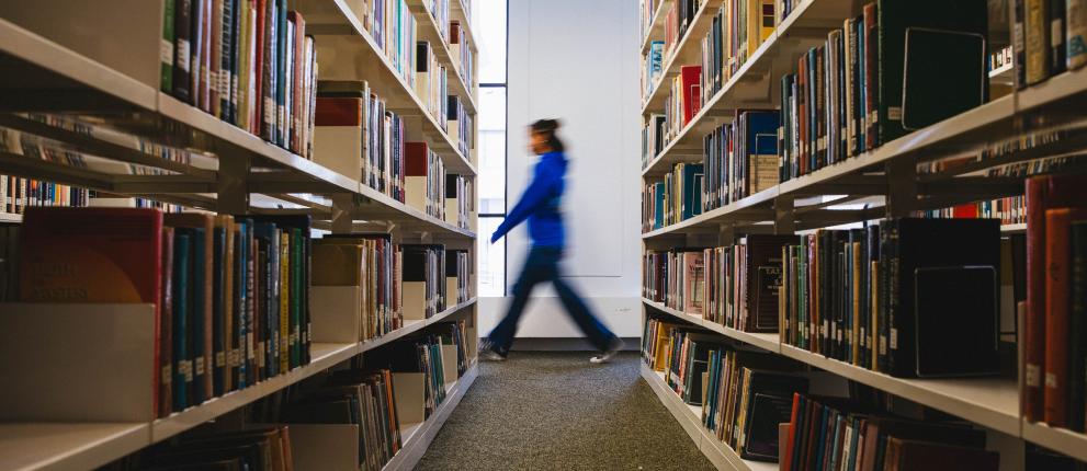 A person in a blue jacket walks past bookshelves filled with books in a brightly lit library aisle.