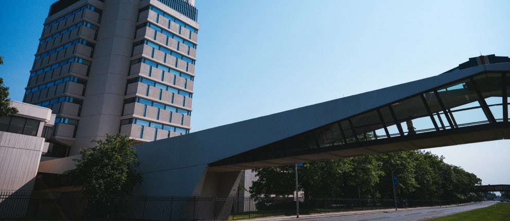 A tall modern building with many windows stands next to a covered skywalk that crosses over a road lined with trees on a clear, sunny day.
