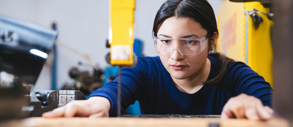 A person wearing safety goggles operates a bandsaw in a workshop, focusing intently while guiding material through the saw blade with both hands.
