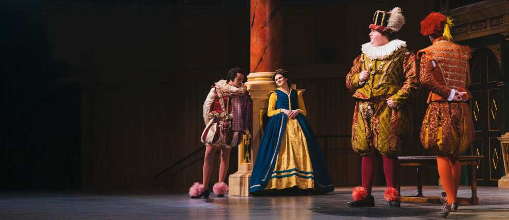 Four actors in colorful, ornate Renaissance costumes perform on stage; one woman in a blue and gold dress leans against a pillar, while three men in elaborate outfits stand and interact beside her.