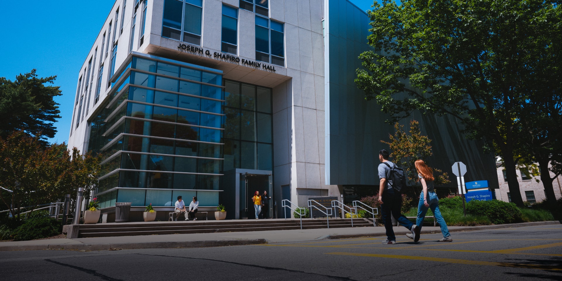 Two students walk past the entrance of the Joseph G. Shapiro Family Hall, a modern building with large glass windows, trees, and people sitting outside on a sunny day.