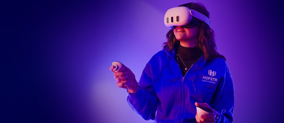 A person wearing a virtual reality headset and holding controllers smiles while standing against a purple background. They are wearing a Hofstra University jacket.