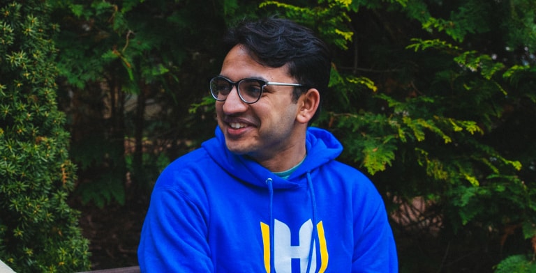A person wearing glasses and a blue hoodie with a large yellow and white Hosftra logo is smiling while sitting outdoors in front of green foliage.