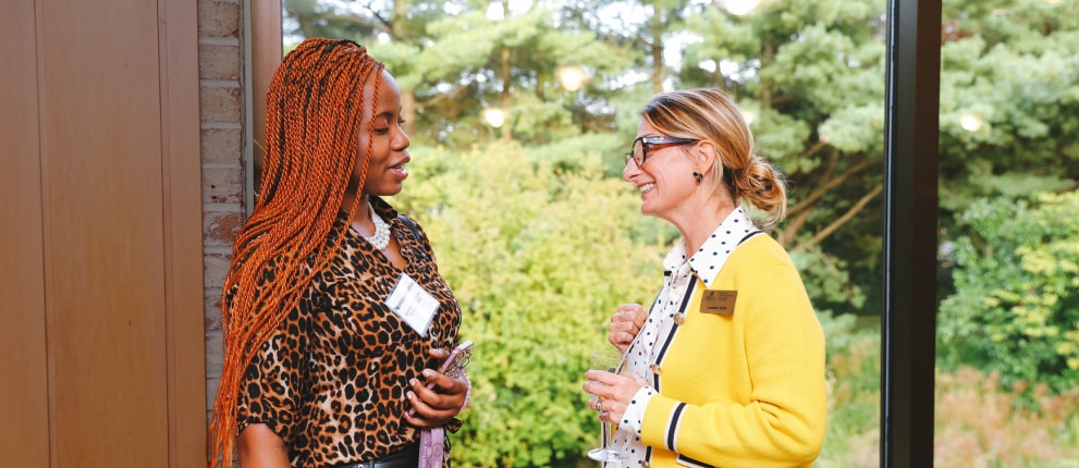 Two women stand indoors by a large window, having a conversation. One wears a leopard print top and has long braids, while the other wears glasses and a yellow cardigan. Green trees are visible outside.