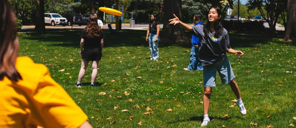 Several young people play frisbee on a grassy lawn surrounded by trees on a sunny day. One person in a gray shirt and shorts is throwing the frisbee, while others watch or prepare to catch it.
