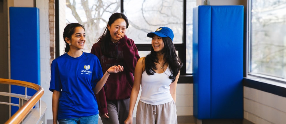Three young women walk and chat in a bright indoor space with large windows. One wears a blue Hofstra University shirt, another a burgundy hoodie, and the third a white tank top and blue cap. They appear happy and relaxed.