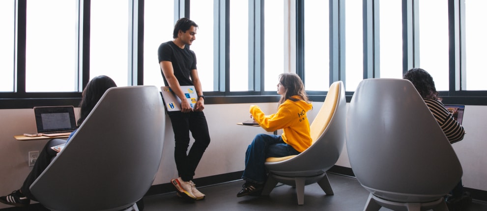 A man holding a laptop stands and talks to a woman in a yellow hoodie seated in a modern chair. Two other people work on laptops in similar chairs by large windows in a bright room.