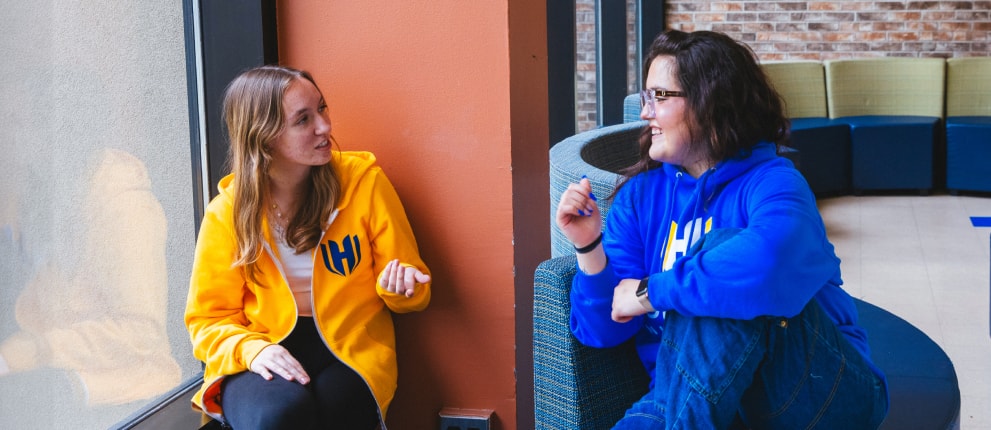 Two people wearing yellow and blue hoodies sit indoors by a window and wall, engaged in conversation and smiling at each other.