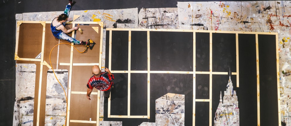 Two people work on a large wooden set or frame on the floor, surrounded by paint-splattered sheets. One uses a tool while kneeling; the other stands nearby, both focused on their tasks. View is from above.