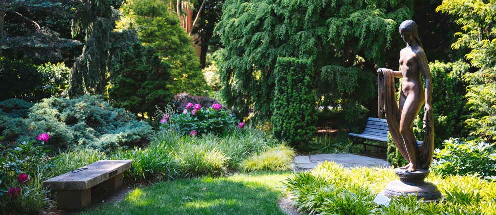 A peaceful garden features a stone bench, blooming flowers, lush greenery, and a bronze statue of a woman standing on a pedestal near a shaded path.