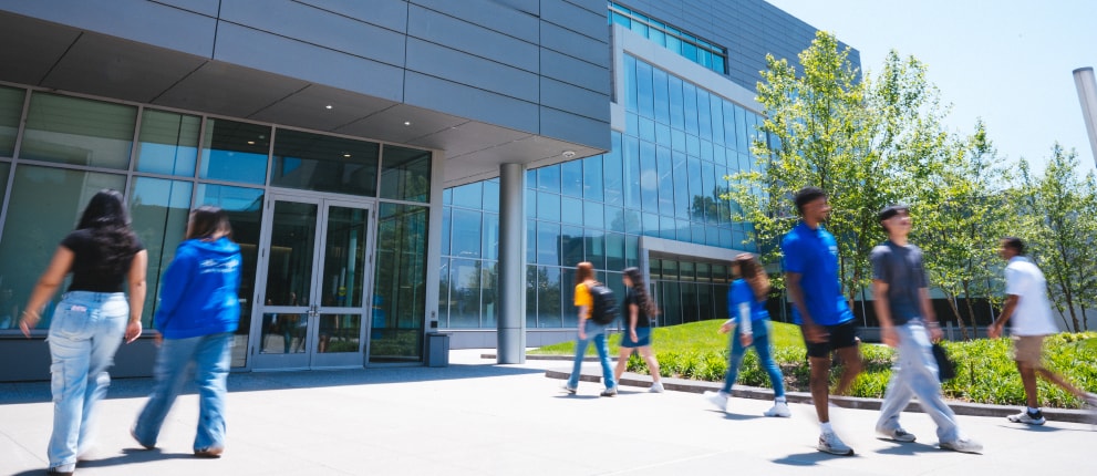 Students walk in front of a modern glass and metal building on a sunny day, with trees and green grass nearby. Some students wear blue university shirts while others wear casual clothes.