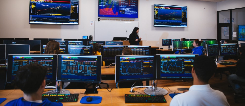 Students sit at desks with multiple monitors displaying financial data in a modern classroom, while an instructor stands at the front near large screens showing charts and graphs.