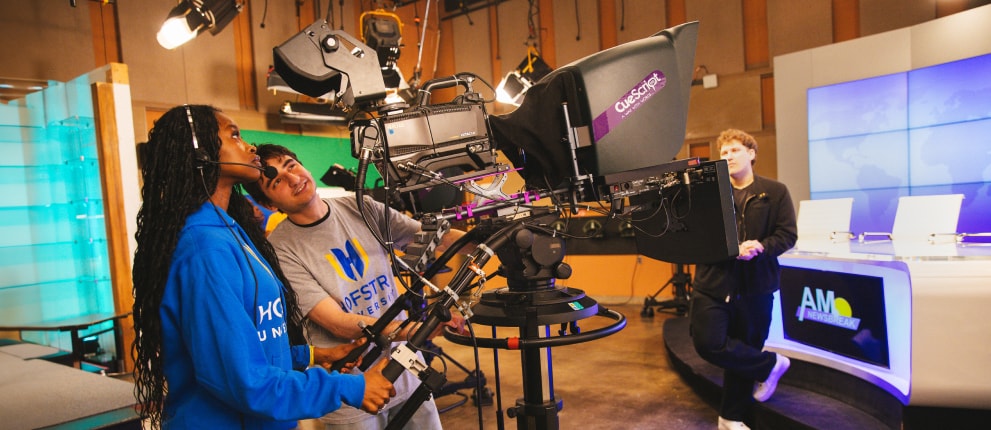 Three people in a TV studio: two operate a large video camera while another stands by the news desk. The studio features bright lights, monitors, and the “AM News” logo on a screen.