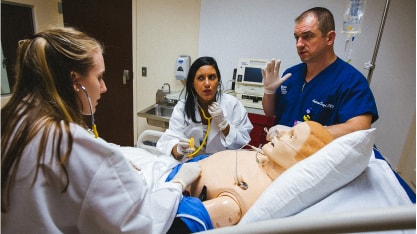 Three medical professionals in scrubs and lab coats practice a procedure on a medical mannequin in a hospital room, using stethoscopes and discussing the simulation.
