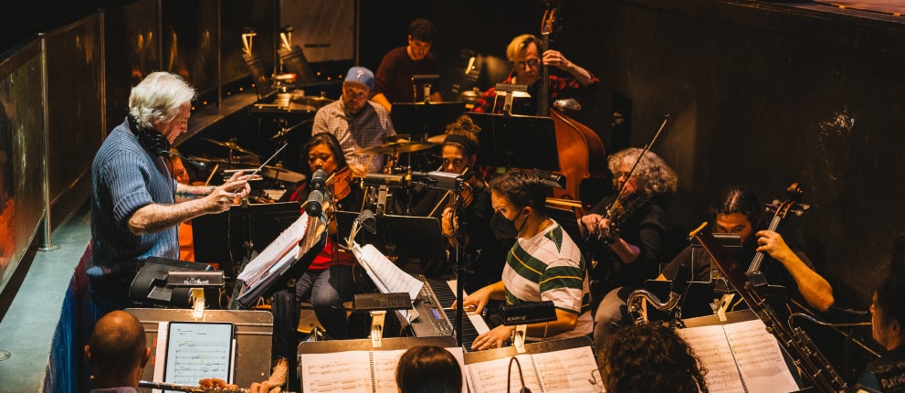 A conductor leads an orchestra in a dimly lit pit, with musicians playing various instruments including strings and brass, all focused on their sheet music.