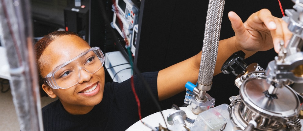 A person wearing safety goggles adjusts scientific equipment in a laboratory, smiling while working with metal pipes and instrumentation.