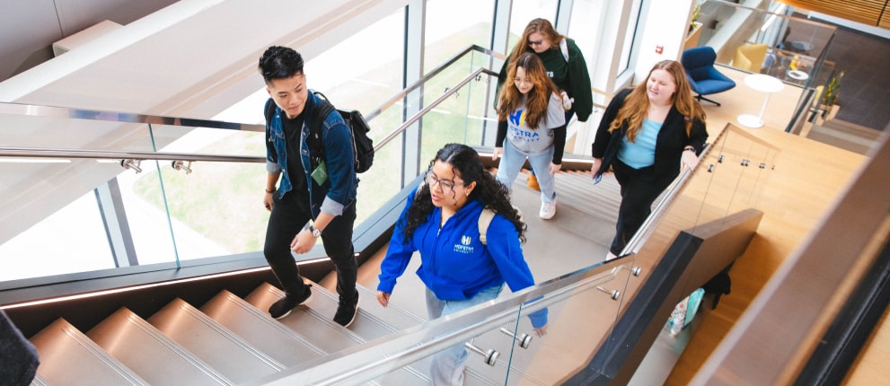 Five students walk up a modern indoor staircase in a bright building, talking and smiling. Some carry backpacks and wear casual clothes, suggesting a school or university setting.