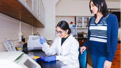 A woman in a lab coat and gloves uses a pipette at a laboratory bench while another woman, wearing a striped sweater, stands beside her, observing and smiling.