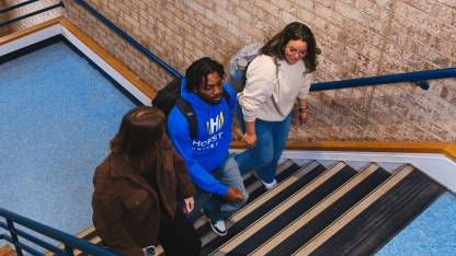 Three students walk up a staircase together in a school building, talking and smiling. The stairs are striped, and the walls are brick. The students carry backpacks and wear casual clothes.