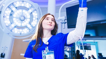 A healthcare professional in blue scrubs and gloves adjusts an IV drip in a brightly lit medical room with surgical lights and monitors in the background.