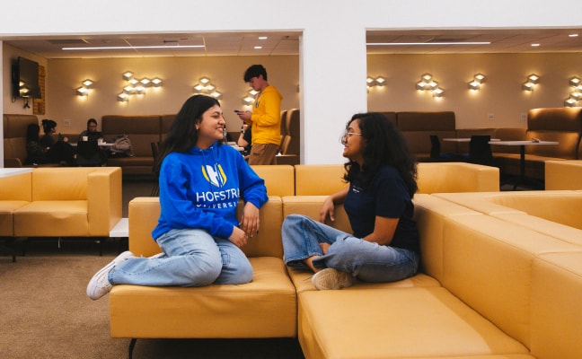 Two students sit on yellow couches in a lounge, smiling and talking. One wears a blue Hofstra University sweatshirt. Other students are in the background, some sitting and one standing near the wall.