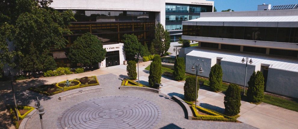 A circular stone plaza with a labyrinth pattern is surrounded by trimmed bushes, trees, and modern academic buildings on a sunny day.