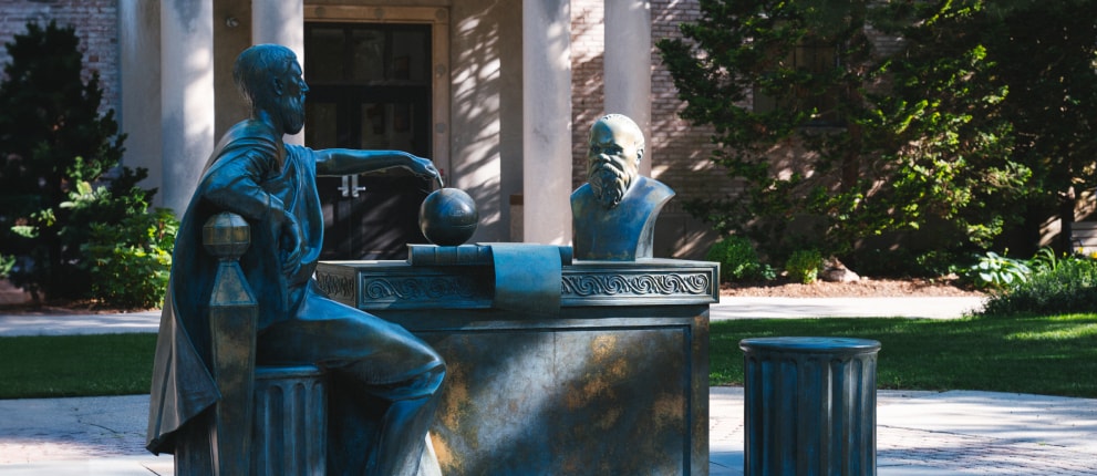 Two bronze statues sit at a table outdoors, one holding a sphere and the other with a hand on its chin in a thinking pose. Trees and a building entrance are in the background.
