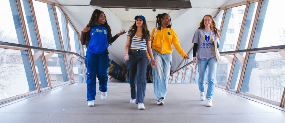 Four young women walk together across a glass-enclosed skybridge, smiling and talking. They wear casual clothes, including jeans, T-shirts, and sweatshirts, with some displaying university logos. Trees and buildings are visible outside.