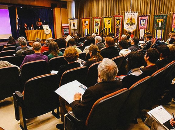 Audience seated in a conference room facing a stage with speakers at a table. Colorful banners with crests hang at the front of the room. People hold papers and listen attentively.