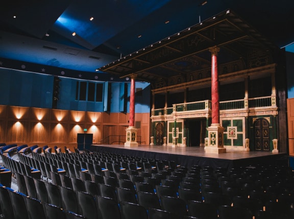 An empty theater auditorium with rows of black seats facing a stage decorated with classical columns, balconies, and ornate details, illuminated by soft overhead lighting.