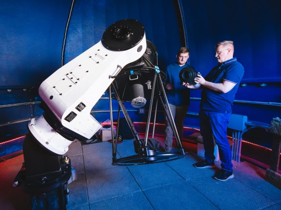 Two people stand inside an observatory, examining parts near a large white telescope on a mount, with a blue domed ceiling in the background.
