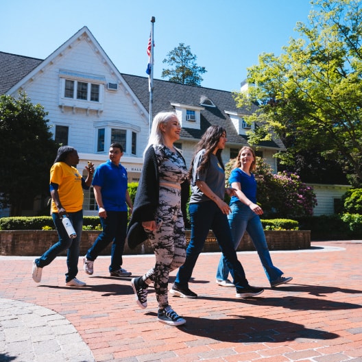 Five young adults walk together on a brick path in front of a large white house on a sunny day, with trees and greenery in the background.