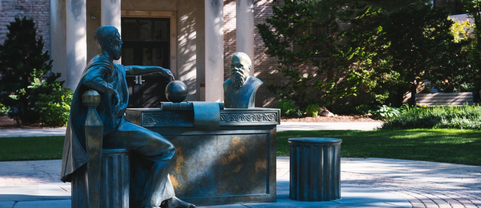 Bronze statues of two figures, one seated at a desk with a globe and another as a bust on the desk, are displayed outdoors in a leafy, sunlit courtyard.
