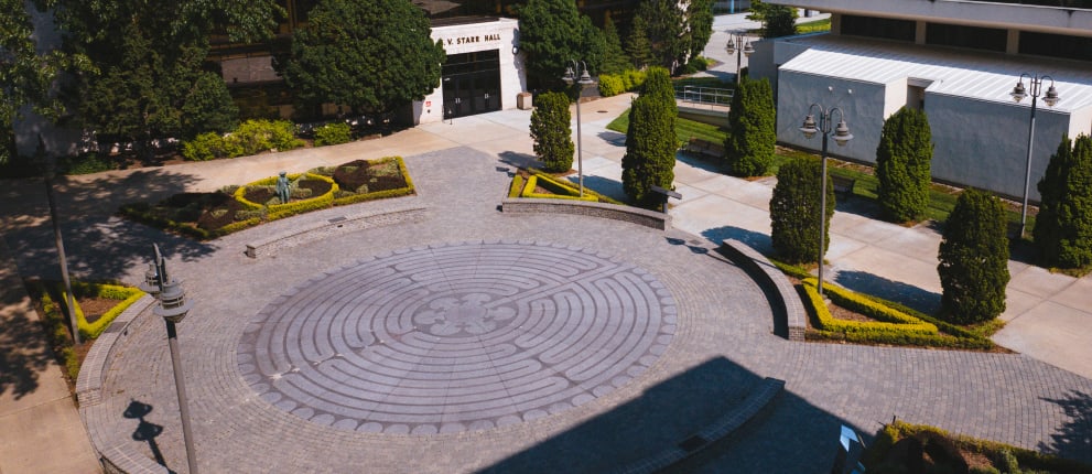A circular brick labyrinth with a cloverleaf pattern is set in a paved plaza, surrounded by manicured shrubs, trees, and nearby campus buildings.