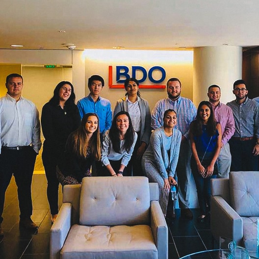 A group of eleven people pose and smile in an office lobby in front of a wall with the BDO logo. Some are standing, while others are sitting or kneeling. The setting looks modern and professional.