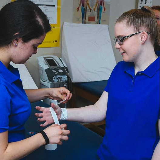 Two women in blue shirts, one wrapping a bandage around the other’s wrist in a clinical setting. Medical posters and equipment are visible in the background.