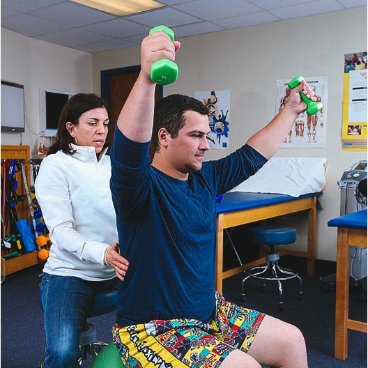 A physical therapist assists a man lifting green dumbbells overhead while seated on an exercise ball in a clinical setting with medical equipment and posters in the background.