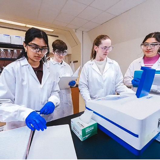 Four students wearing lab coats, gloves, and safety glasses work together in a laboratory, using scientific instruments and taking notes at a workstation.