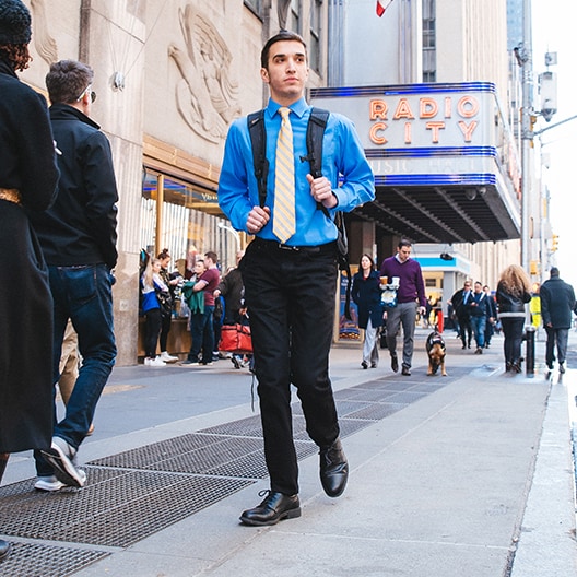A man in a blue shirt, black pants, and a yellow tie walks with a backpack near Radio City Music Hall in New York City, surrounded by people on a busy sidewalk.