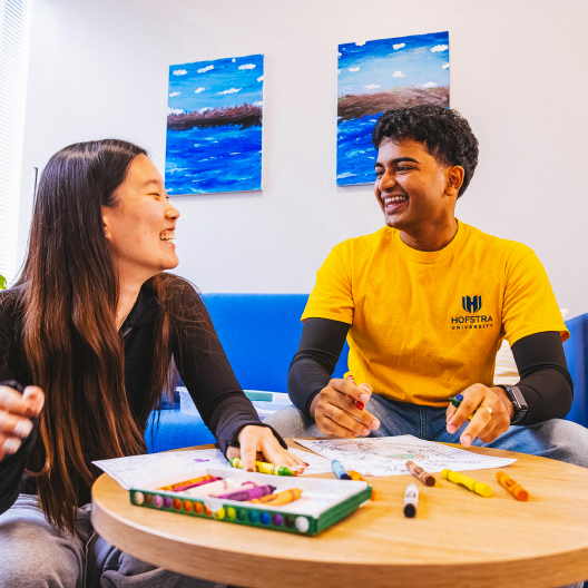 Two students sit at a round table coloring with markers and colored pencils, smiling at each other. One wears a yellow Hofstra University shirt. There are paintings of the ocean on the wall behind them.