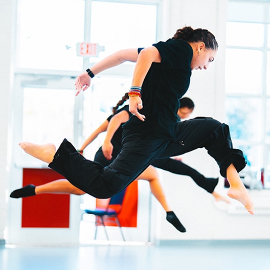 A person dressed in black jumps energetically in a bright room, with another dancer blurred in the background. Both appear to be mid-dance, showcasing dynamic movement and athleticism.