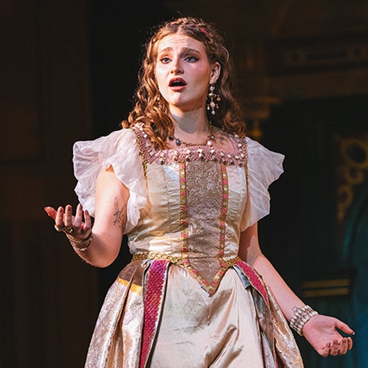 A woman in a gold and white Renaissance-style dress sings on stage, raising one hand expressively. She has curly hair, wears decorative jewelry, and appears to be performing in a theatrical or operatic production.
