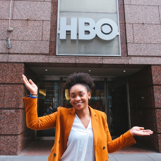 A smiling person in a mustard blazer stands outside a building with large HBO letters above the entrance, arms raised in excitement.