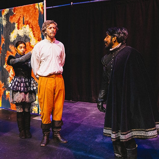 Three actors perform on stage; one man stands with hands behind his back, another man in a dark cape faces him, and a woman in a patterned dress stands behind, holding the first mans arms. A colorful backdrop is partially visible.