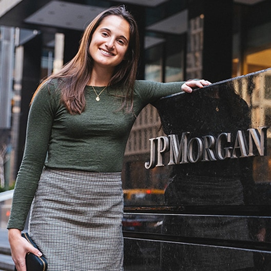 A woman with long brown hair, wearing a green top and plaid skirt, smiles while standing beside a black JP Morgan sign outside an office building.