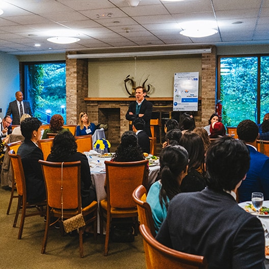 A person stands and speaks to a group of people seated around round tables at a formal indoor event; food and drinks are on the tables, and large windows show greenery outside.