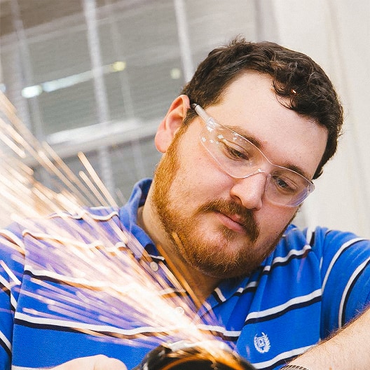 A man wearing safety glasses and a blue striped shirt operates a power tool, creating bright sparks as he works.